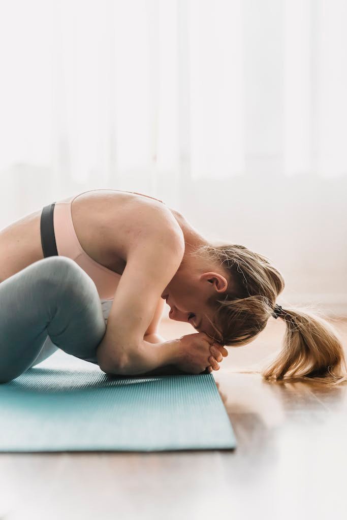 Side view of focused young fit woman with long blond hair in sportswear stretching thighs while doing Baddha Konasana B yoga pose with closed eyes in sunny studio