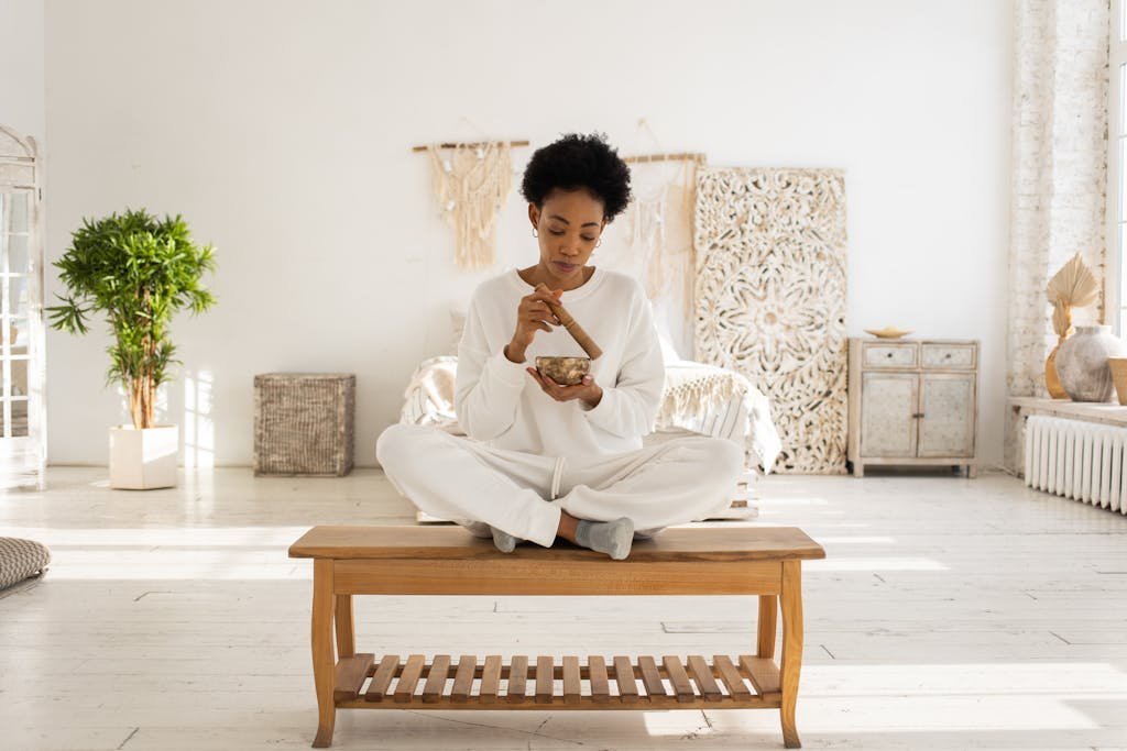 Woman meditates with a singing bowl in a cozy, light-filled bedroom, sitting cross-legged on a wooden bench.