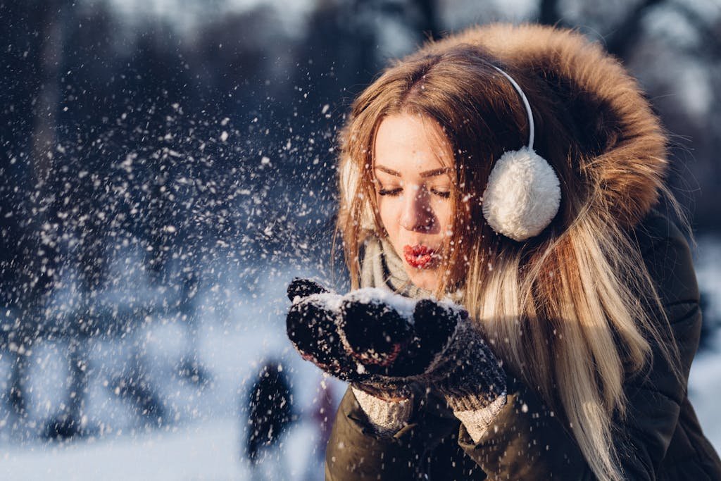 A young woman enjoying a snowy day, blowing snow while wearing cozy earmuffs and a winter coat.