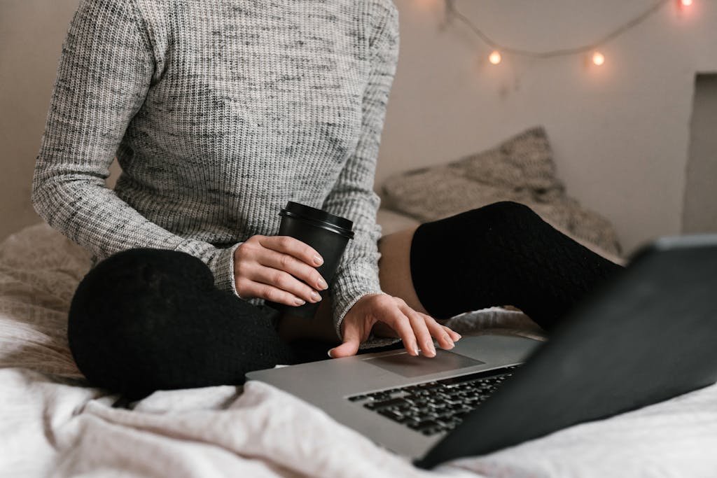 Woman in sweater with coffee and laptop on bed, perfect for relaxed indoor work or study settings.
